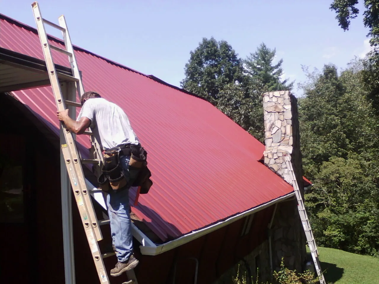 Roofer on a ladder beside a red metal roof and stone chimney — Soffit & Fascia Repair by Roofing Brothers in Weehawken